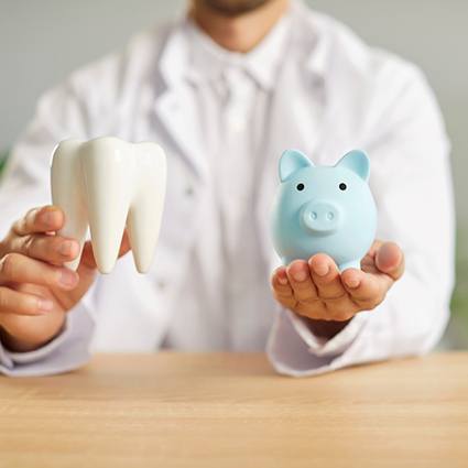 Man in white jacket holding model tooth in one hand and blue piggy bank in the other