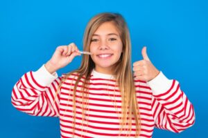 Teenager smiling and holding clear aligner while giving thumbs up.
